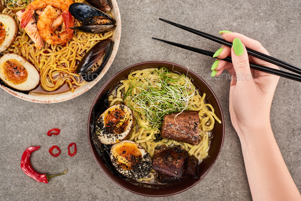 cropped view of woman holding chopsticks near spicy seafood and meat ramen on grey surface - Stock Photo - Images