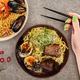 cropped view of woman holding chopsticks near spicy seafood and meat ramen on grey surface - PhotoDune Item for Sale