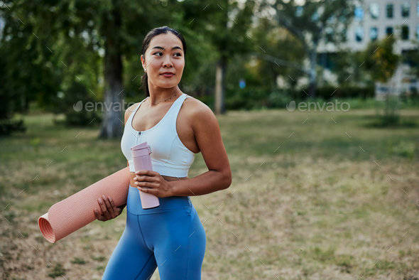 Fit sportswoman on the way to yoga class outdoors, holding yoga mat and  bottle of water Stock Photo by bernardbodo