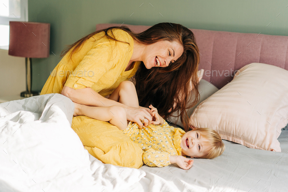 Young laughing mother tickles baby daughter in bed.