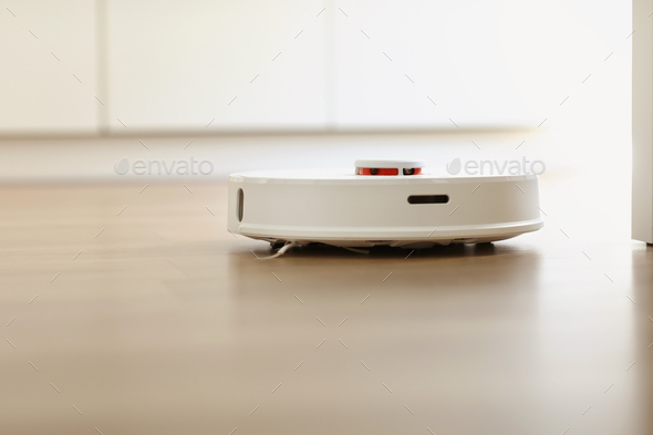 White robotic vacuum cleaner on laminate floor cleaning dust in living room interior. Smart