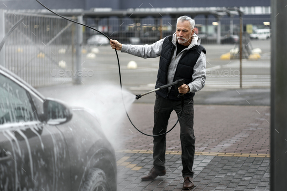 Elegant stylish senior man cleaning car with water gun on self-service washing station. Full-length
