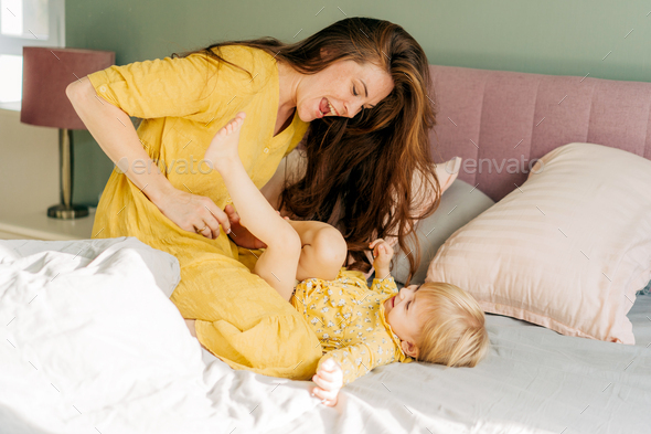 Young laughing mother tickles playing baby daughter in bed.