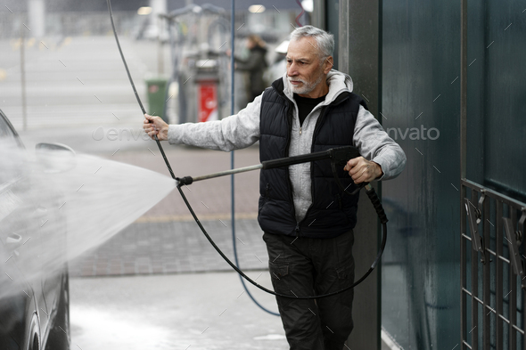Elegant stylish gray-haired senior man cleaning car with a water gun on self-service washing station