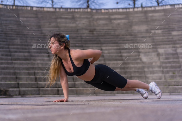 Caucasian young woman doing fitness in city park, with one hand isometric crunches or stabilization