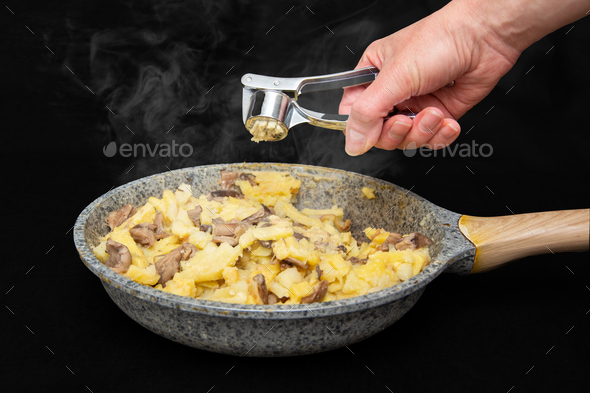 Female hand crushes garlic through juicer in a fried potato with mushrooms in a gray with spots pan