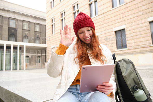 Happy redhead girl, student sits with backpack and digital tablet on street, says hello and waves