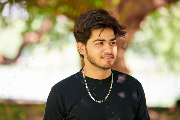 Attractive young indian man portrait in black t shirt and silver neck chain on green park background