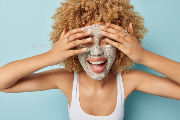 Happy curly haired woman tries to hide keeps hands over face applies clay facial mask to reduce