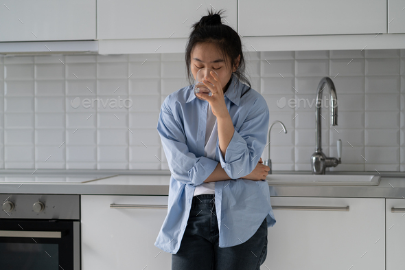 Stressed young Asian woman standing in kitchen drinking water calming down after panic attack