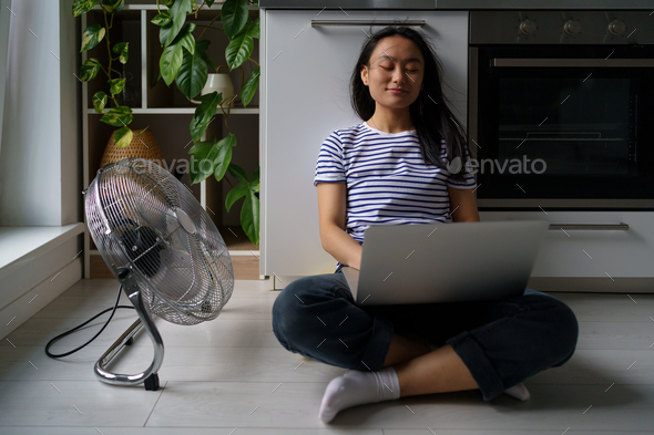 Contented Asian woman turn on floor ventilator fan while work with laptop joyful self save from heat