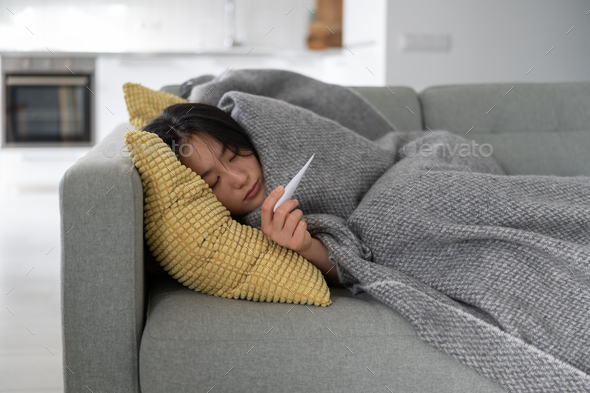 Young sick unhealthy Asian woman suffering from fever measuring temperature while lying on sofa