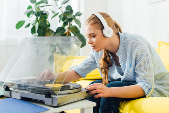 Side view of attractive girl in headphones using record player on coffee table at home