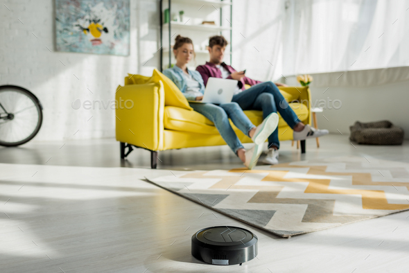selective focus of robotic vacuum cleaner washing carpet near man watching movie and woman using