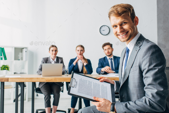 Selective focus of smiling employee with resume looking at camera near recruiters in office