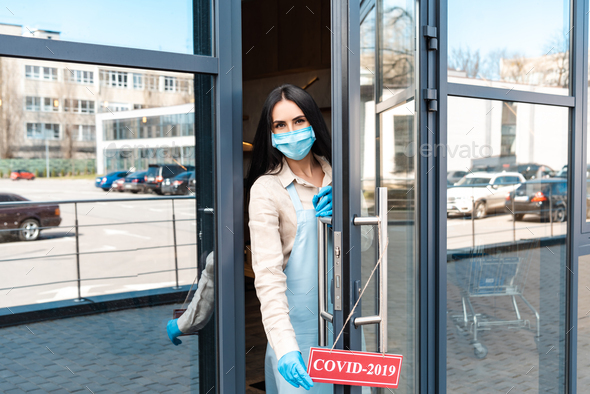 Cafe owner in medical mask near door looking at camera and touching card with covid-2019 lettering