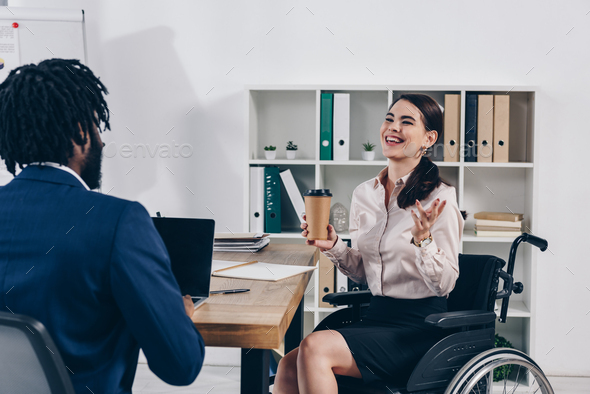 Disabled employee on wheelchair with paper cup of coffee laughing near african american recruiter