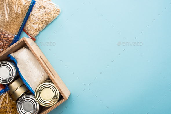 top view of cans and groats in zipper bags in wooden box on blue background, food donation concept