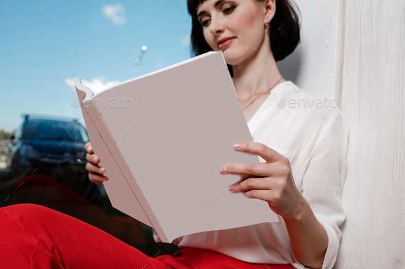 beautiful young woman holding a magazine with mock up. girl in a white shirt and red pants sitting
