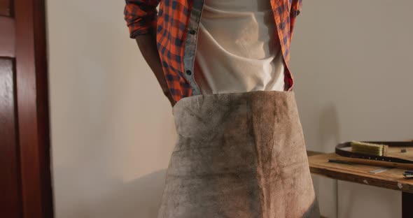 Midsection of african american craftsman putting apron on in leather workshop alt