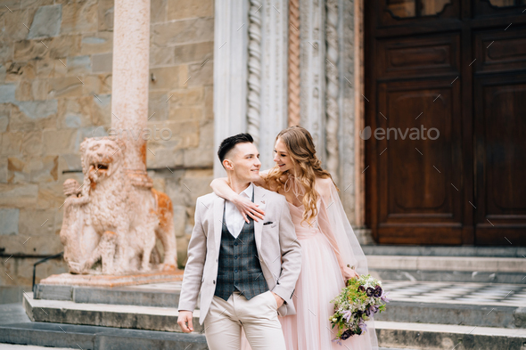 Bride with a bouquet of flowers hugs shoulders of smiling groom on the steps at the entrance to the