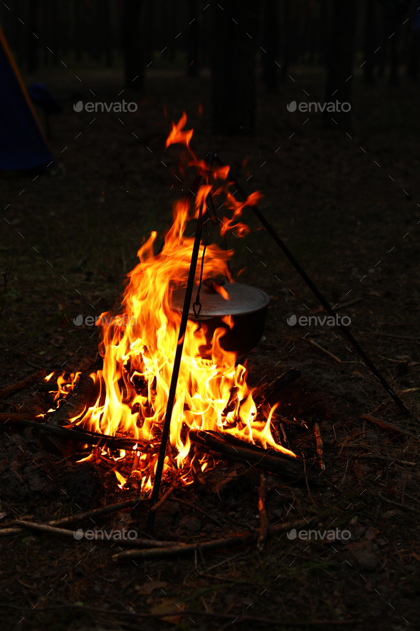 Black large tourist pot hangs over burning flame of campfire in the forest.  Cooking dinner on