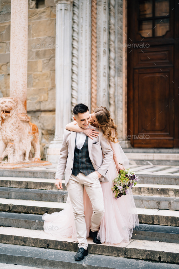 Bride with a bouquet of flowers hugs from behind smiling groom on the steps at the entrance to the