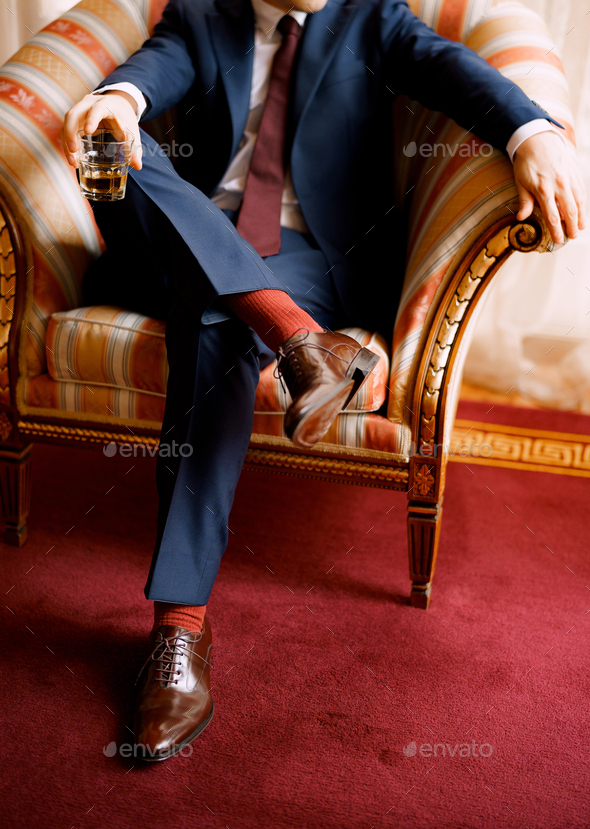 Legs of a man in blue pants, red socks and brown shoes sitting in the chair  holding glass in one Stock Photo by Nadtochii