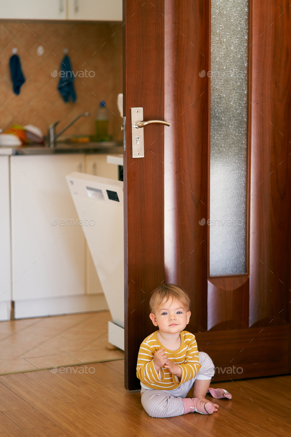 Cute kid in a striped blouse sits on the kitchen floor against the background of an open door and a