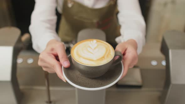 Close up of Asian barista woman holding latte coffee artwork hot cup giving to customer in cafe. alt