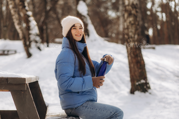 Woman drinking hot tea holding vacuum flask in winter park.