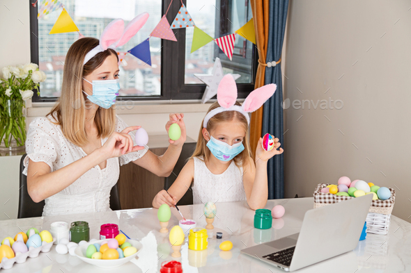 Pretty little girl wearing bunny ears and disposable medical face mask and painting easter eggs at