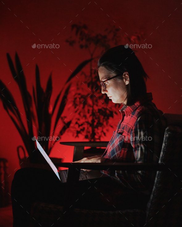 woman is working on laptop in home office in evening. red lamp in cozy interior - Stock Photo - Images