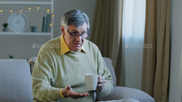 Old elderly mature grandfather man sitting at home couch watching TV drinking hot tea from cup