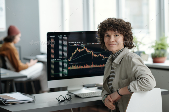 Young smiling female analyst sitting by workplace with computer monitor