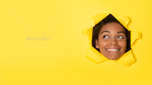 Happy black lady posing in hole in torn paper looking aside, advertising great offer over yellow