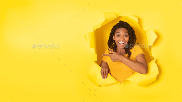 Excited black woman pointing finger aside at free space, posing in hole in torn yellow paper