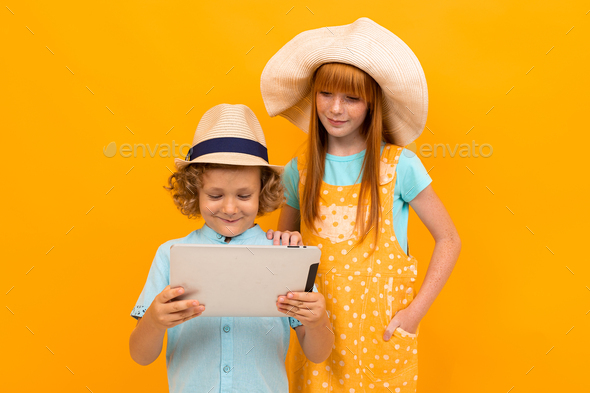 Red-haired brother and sister in summer hats book tickets on a tablet on a yellow background.