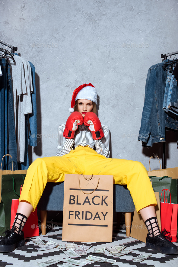 girl in boxing gloves and santa hat sitting with shopping bags and clothes around, black friday