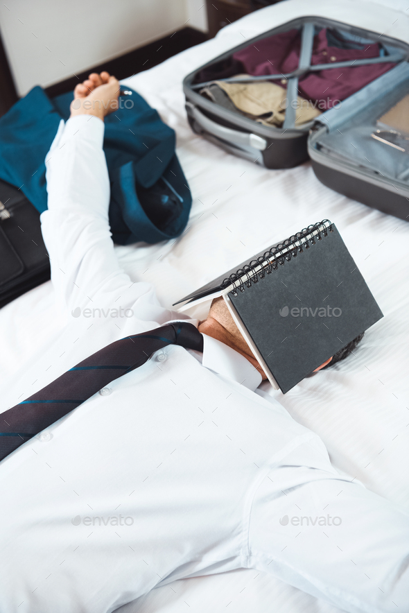 Man in formal wear lying on bed in hotel room with notebook on his face