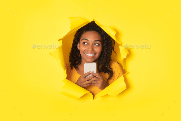 Excited black lady posing with cellphone and looking aside at free space in torn hole in yellow