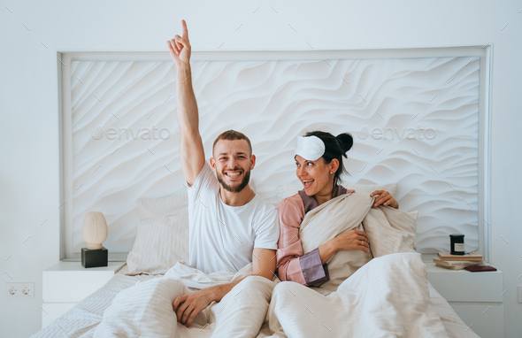 Happy young beardy man sits on bed with wife raises hand points up by index finger. Awaking couple