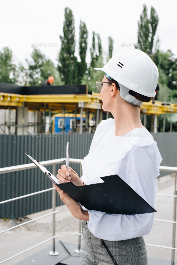 Young businesswoman in hardhat standing with folder and pen, while watching work at construction