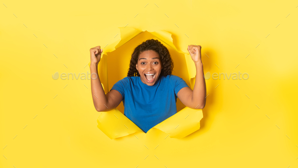 Overjoyed black woman cheering with clenched fists, celebrating victory, posing in torn hole of