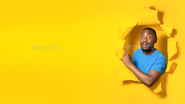 Shocked black man pointing finger aside at free space for design, posing in hole in torn of yellow