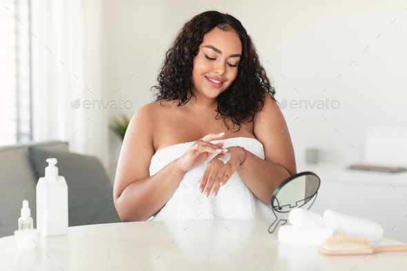 Skincare cosmetics concept. African american plus size lady applying hands cream, sitting at