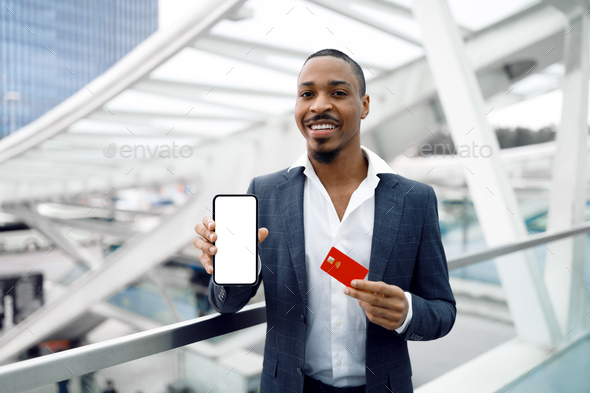 Black Businessman Showing Smartphone With Blank Screen And Credit Card In Airport