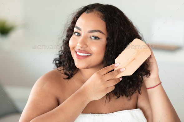 Portrait of happy body positive lady brushing her curly dark hair and smiling at camera, home