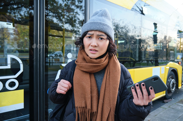 Portrait of confused asian girl, standing on bus stop, holding mobile phone, looking shocked and