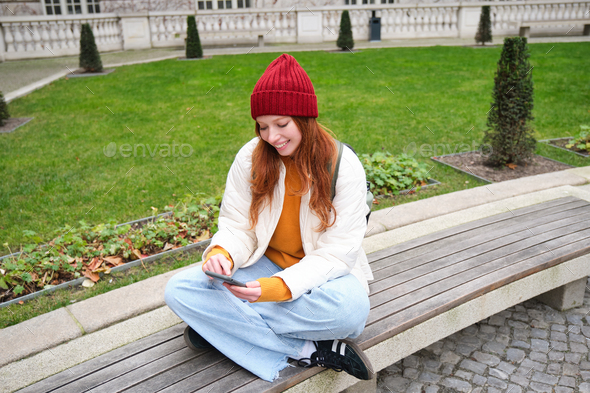 Portrait of stylish young woman, 25 years, sits on bench in park and uses mobile phone, reads